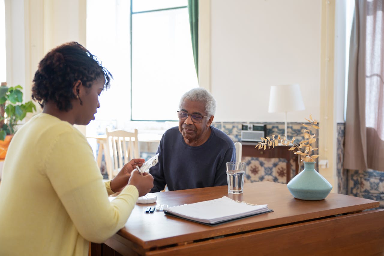 services-01 A caregiver assists an elderly man with medication in a warm, sunlit living room.